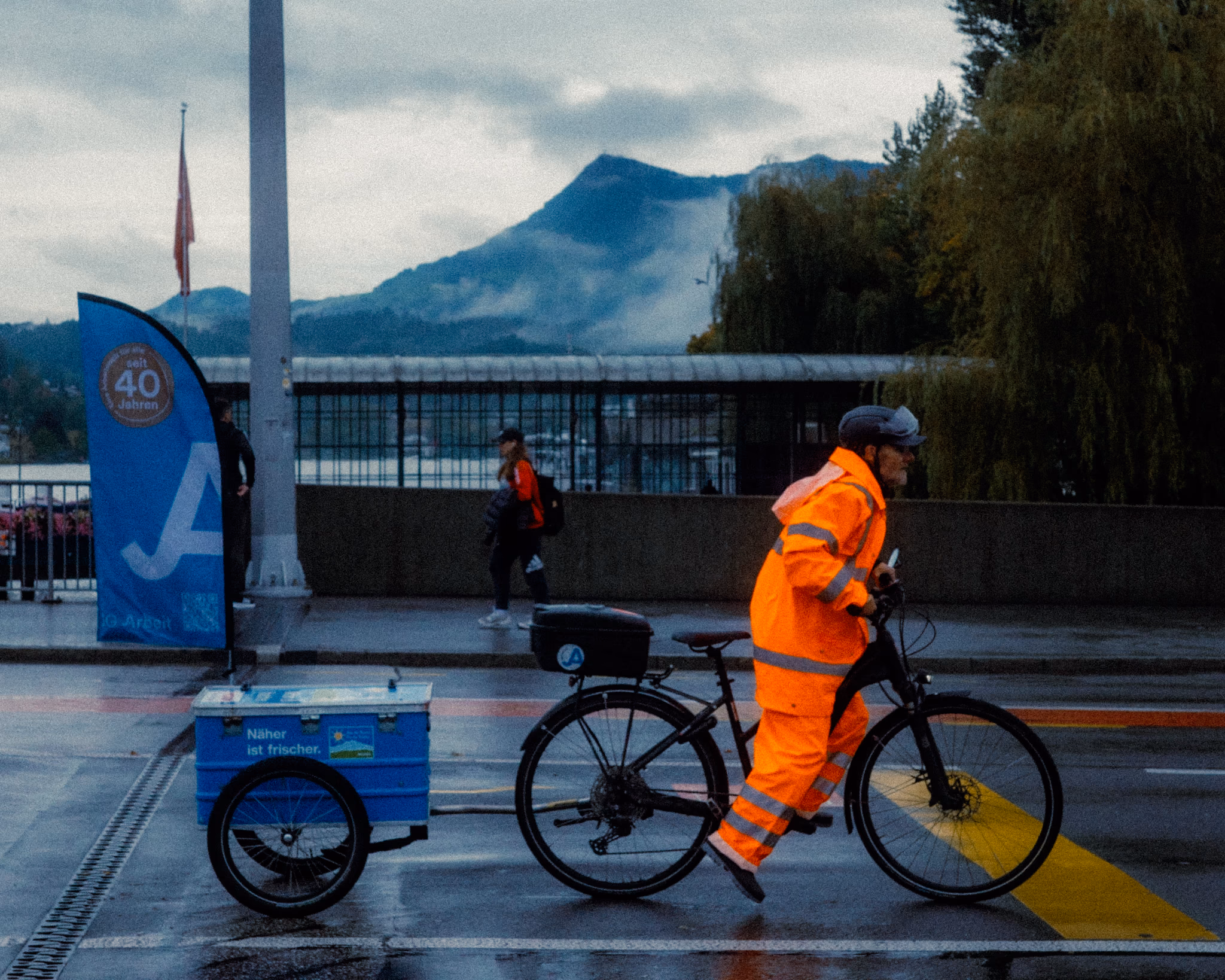 City worker in orange safety vest riding cargo bicycle against misty mountain backdrop at dawn.