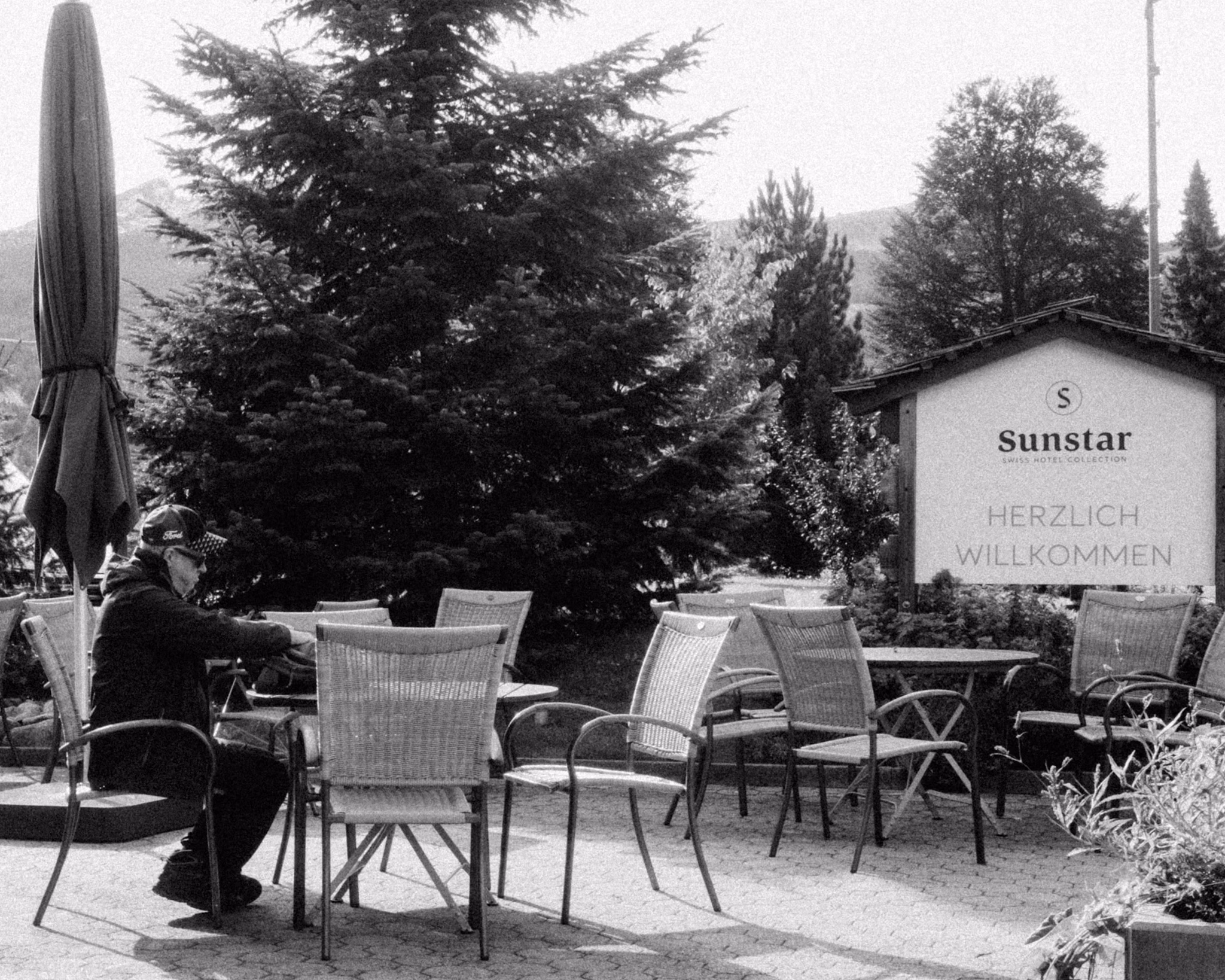 Elderly man sitting alone at outdoor café table with Sunstar hotel sign and tall trees in background, black and white photograph.