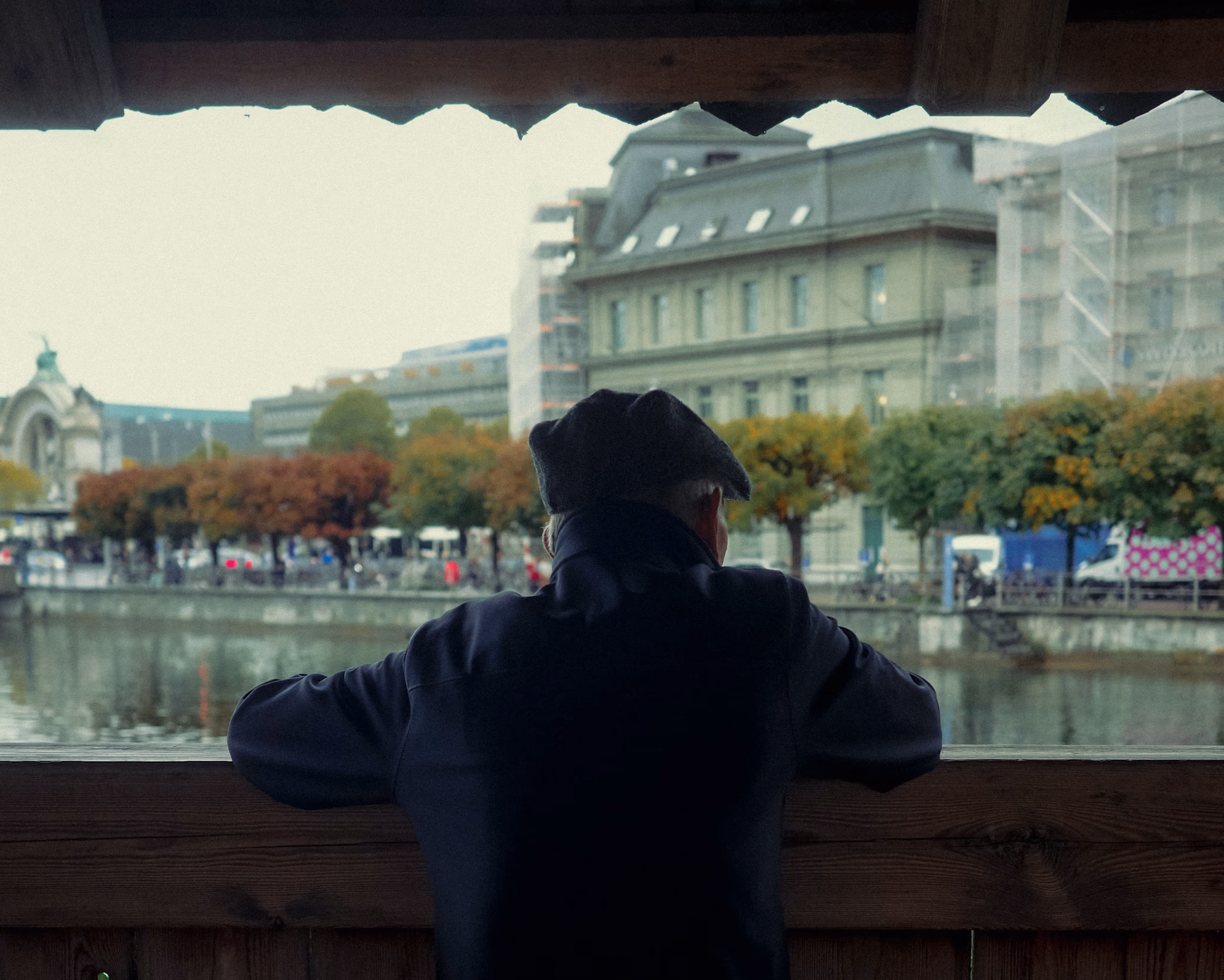 Elderly man in dark coat and beret gazing at autumn cityscape with river and historic buildings.